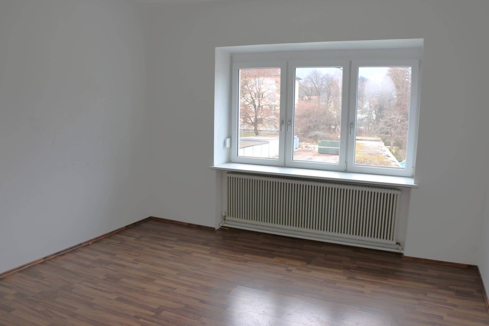 Geräumiges Zimmer mit dunklem Laminatboden und Blick ins Grüne durch das Fenster.