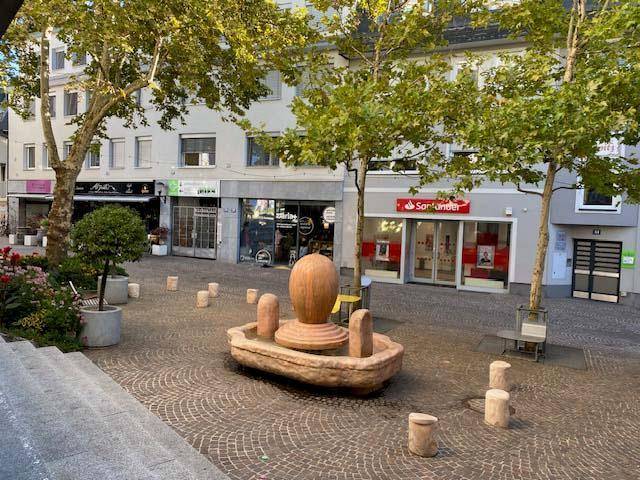 Öffentlicher Platz mit Bäumen, Pflastersteinen und Geschäften im Hintergrund.