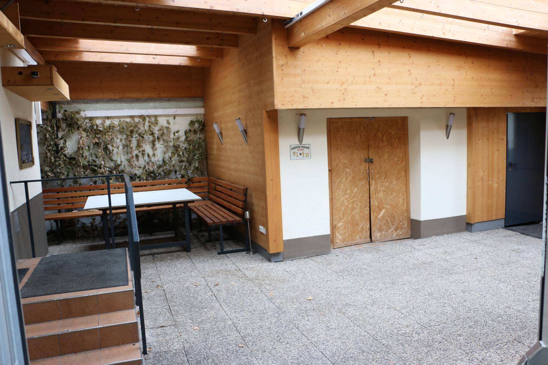 Überdachte Loggia mit Sitzgelegenheiten, Holzbänken und gepflastertem Boden.