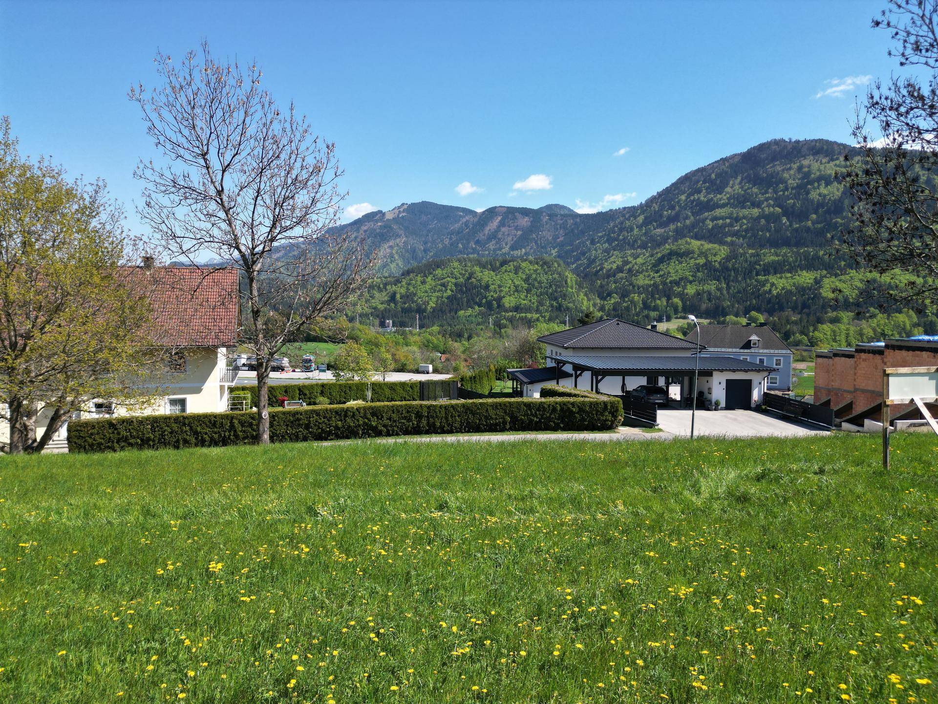 Grünes Feld mit einer Hecke im Vordergrund, dahinter Häuser und eine beeindruckende Berglandschaft.