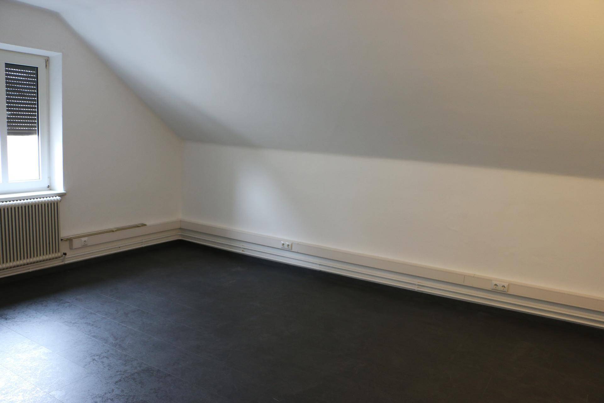 Dachzimmer mit schräger Decke, dunklem Boden und Fenster mit Rollladen.