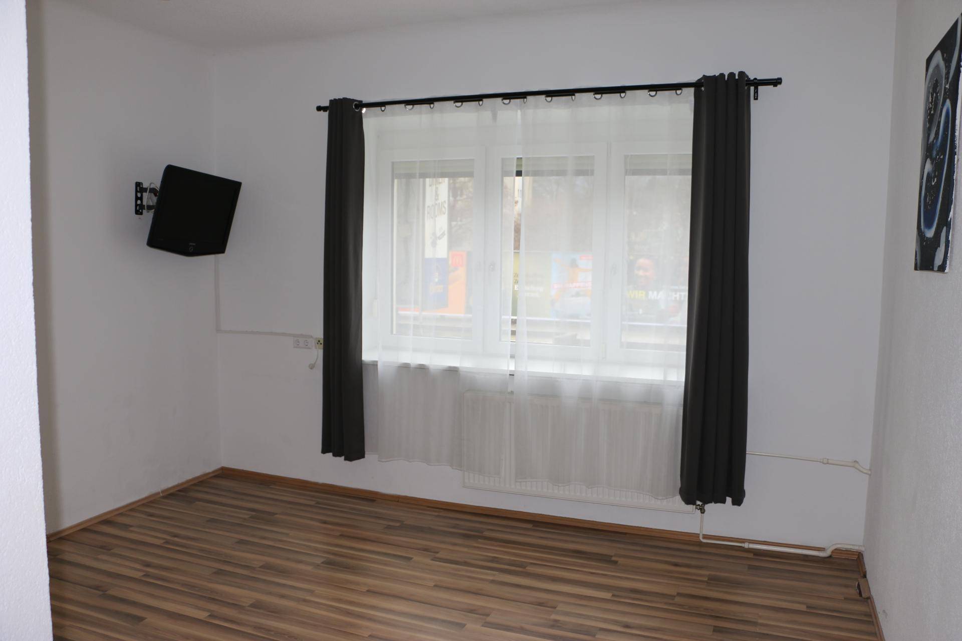 Helles Zimmer mit Laminatboden, großem Fenster und dunklen Vorhängen.