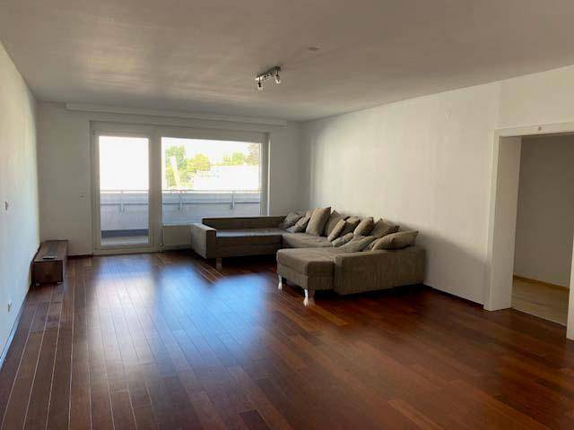 Helles Wohnzimmer mit dunklem Parkettboden, großem Ecksofa und Zugang zum Balkon.