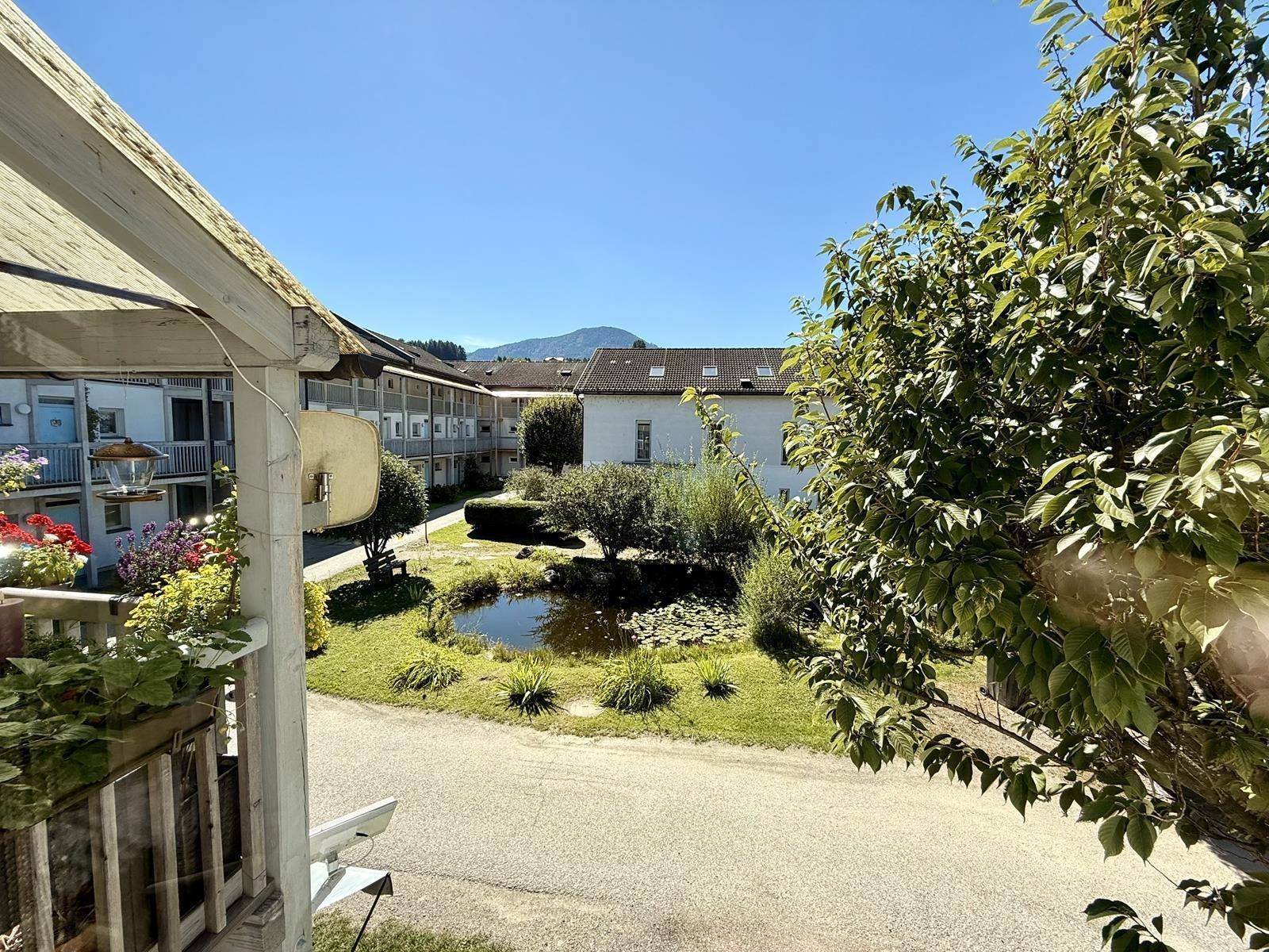 Blick von der Loggia auf einen grünen Innenhof mit Teich und umliegenden Gebäuden sowie Berge im Hintergrund.