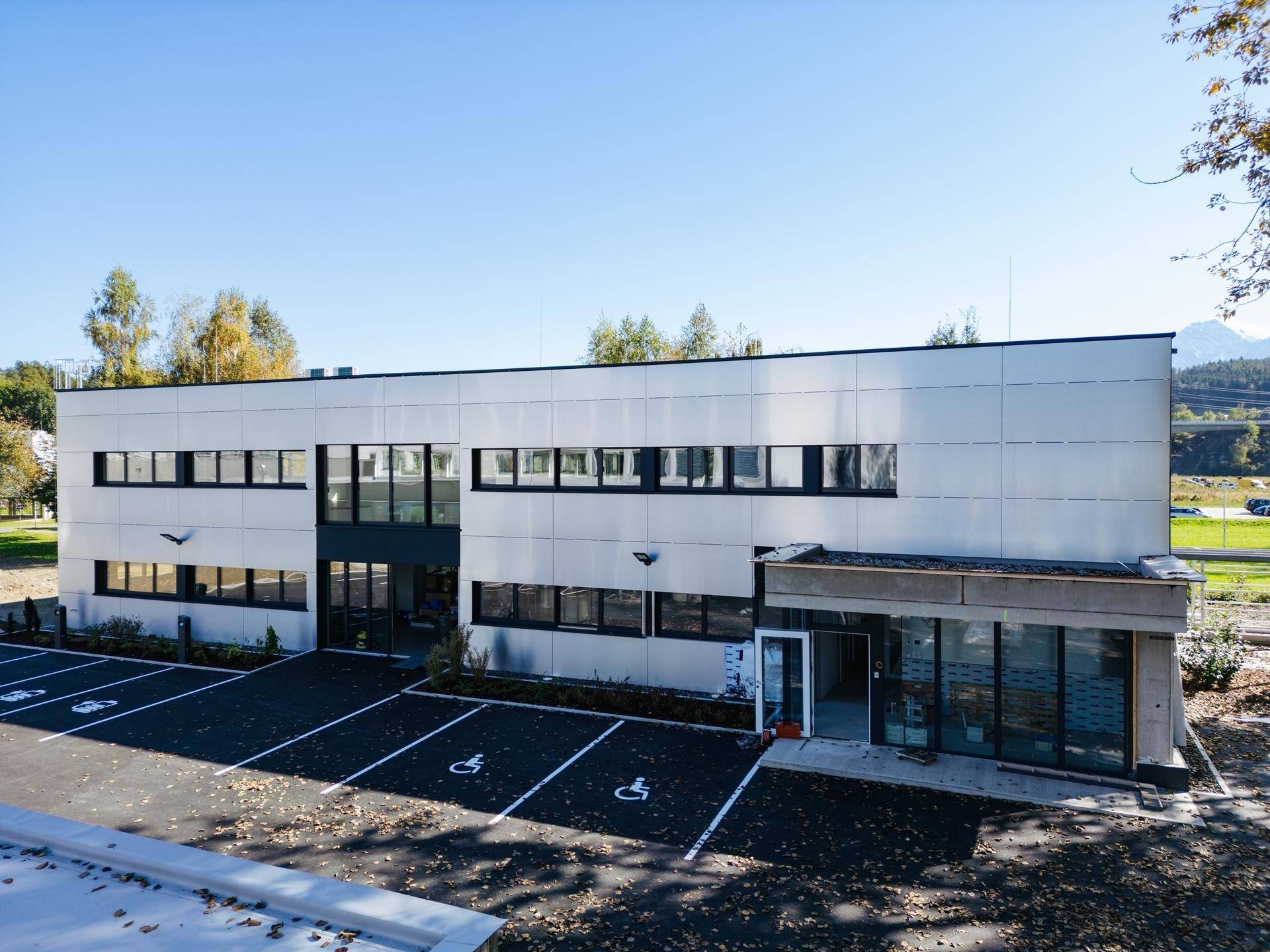 Modernes Geschäftsgebäude mit heller Fassade und barrierefreien Parkplätzen in grüner Umgebung.