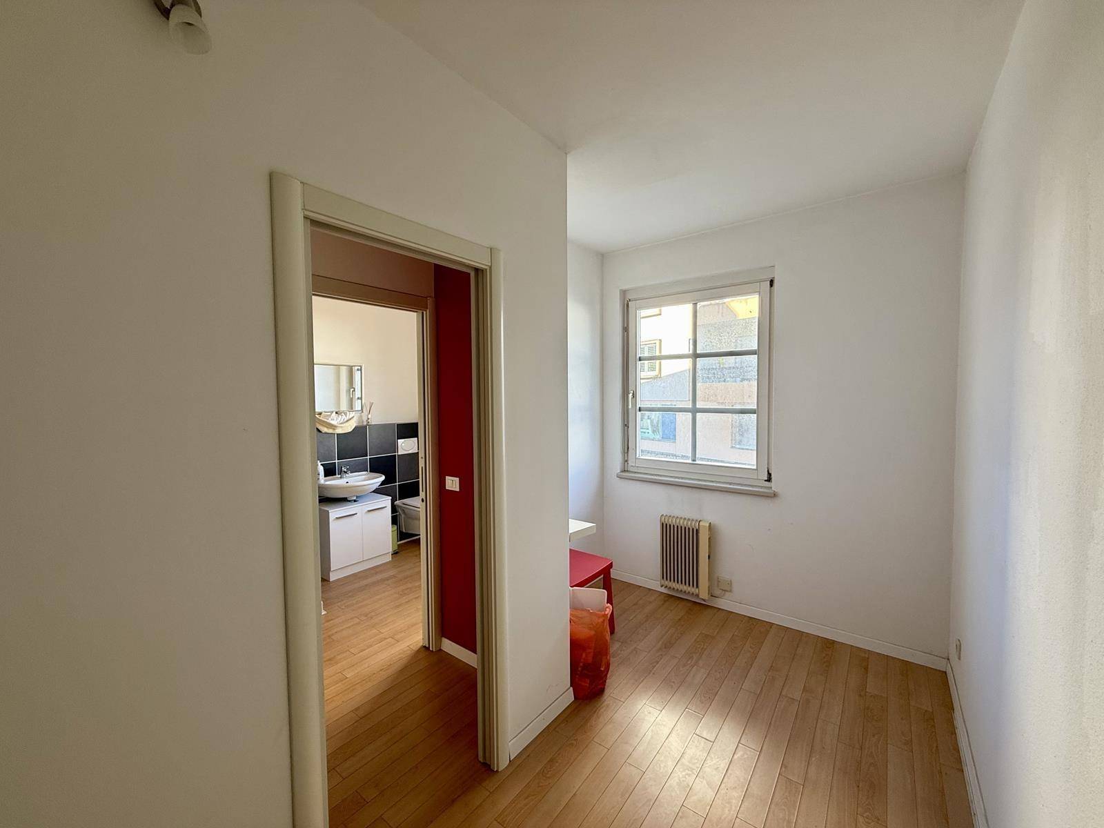 Kleiner Raum mit Holzboden, Fenster und Zugang zu einem weiteren Zimmer, ideal als privater Arbeitsbereich.