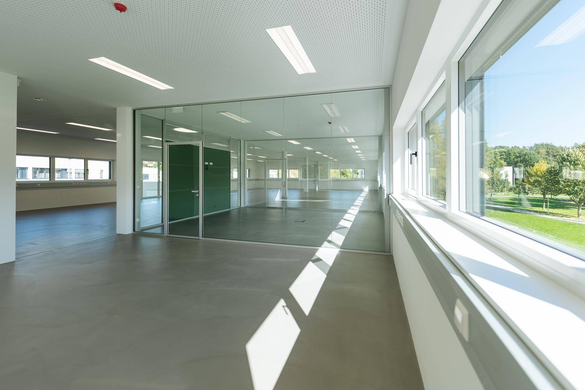 Moderner Büroraum mit Glaswänden, Betonboden und viel Tageslicht durch große Fenster.