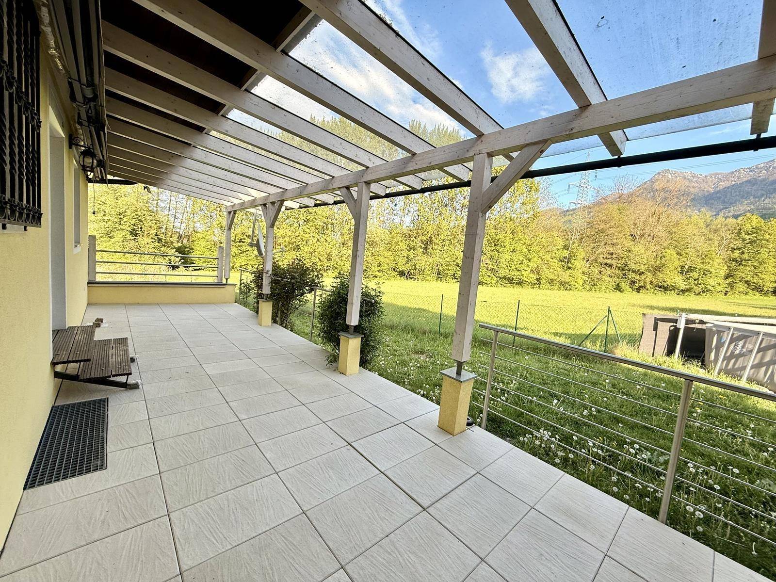 Überdachte Terrasse mit Fliesenboden und Holzpergola, umgeben von Grün und Bergblick.