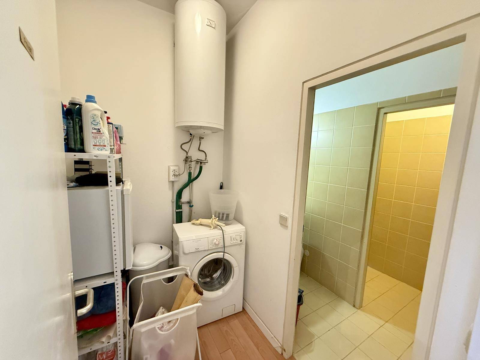 Praktischer Waschraum mit Waschmaschine, Warmwasserboiler und Zugang zu einem weiteren Badezimmer, funktional eingerichtet.