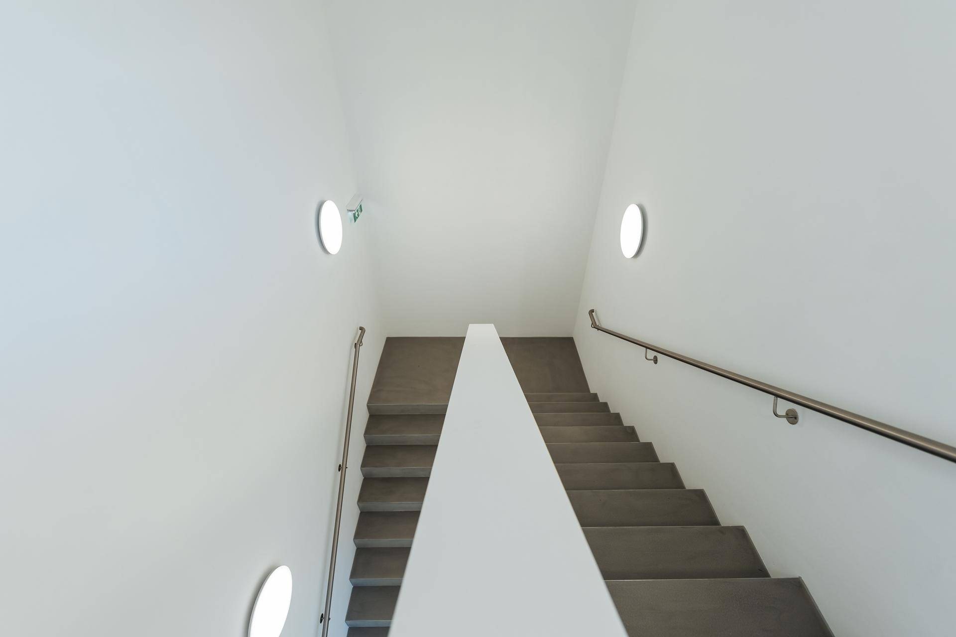 Helles, modernes Treppenhaus mit schlichten Stufen, Handlauf und runden Wandleuchten.