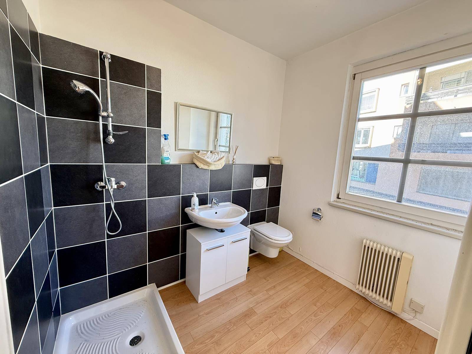 Modernes Badezimmer mit dunklen Wandfliesen, Dusche, Waschbecken und Fenster für natürliche Belüftung und Helligkeit.