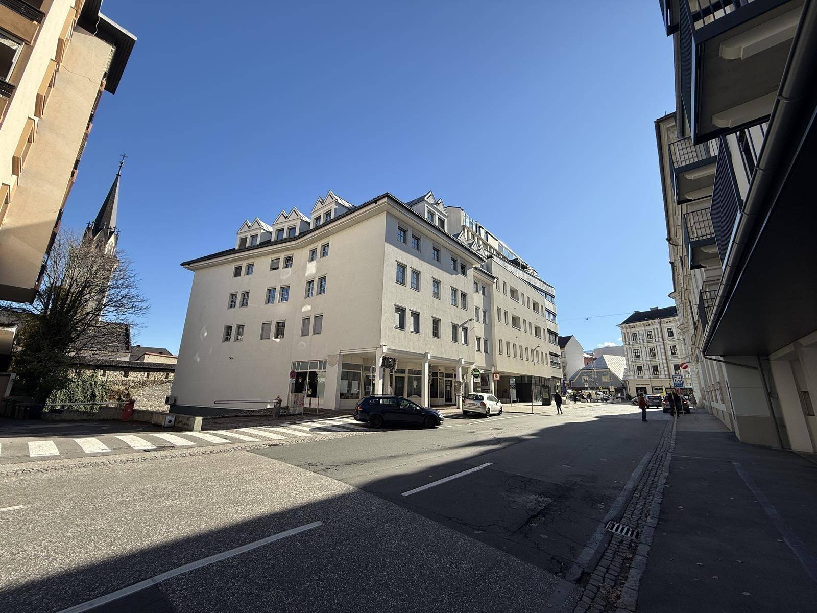 Mehrstöckiges Geschäftsgebäude an einer städtischen Straße mit geparkten Autos und Fußgängern, unter klarem Himmel.