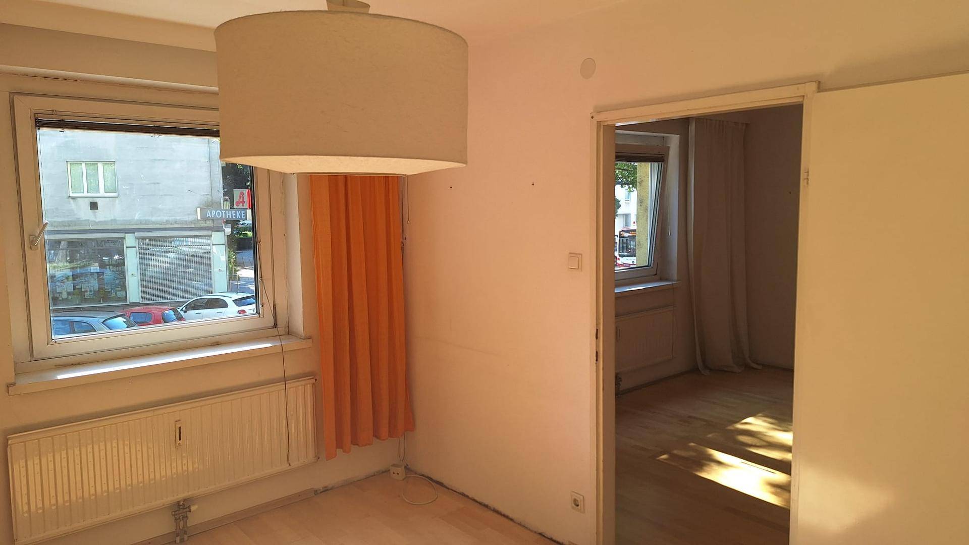 Wohnzimmer mit Parkettboden, großem Fenster und Blick auf die Straße.