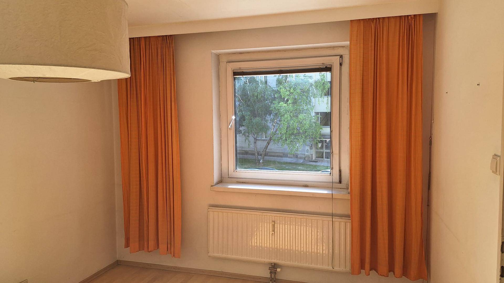 Helles Zimmer mit Parkettboden, Heizkörper und orangefarbenen Vorhängen.