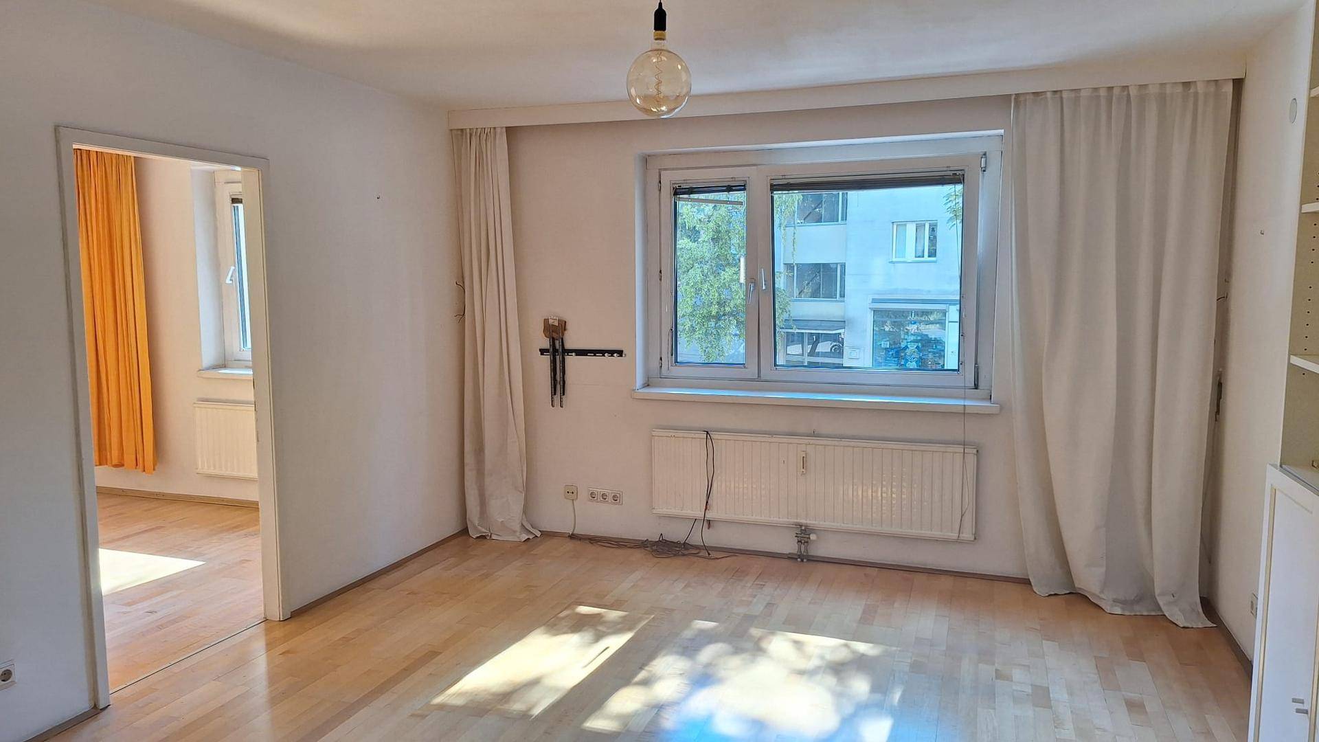 Helles Wohnzimmer mit Parkettboden, großem Fenster und Blick auf die Straße.