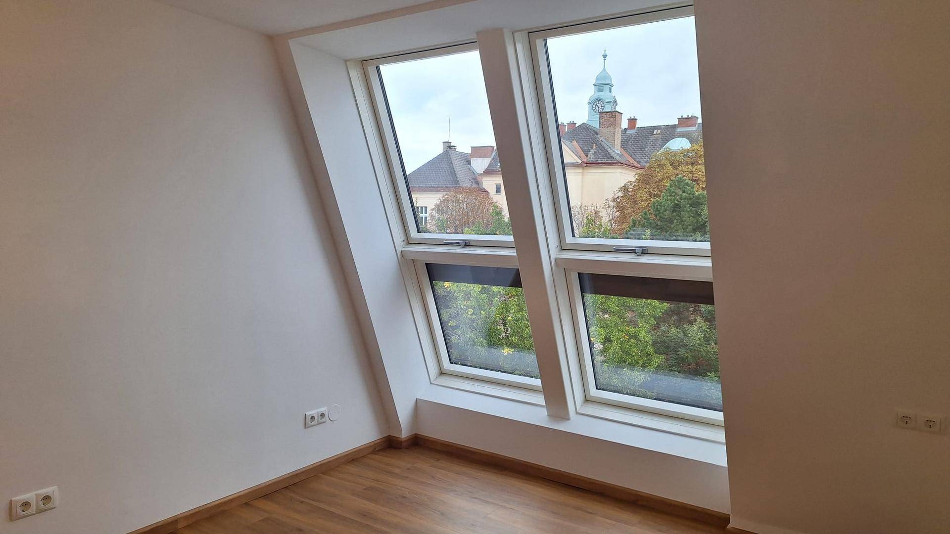 Helles Zimmer im Dachgeschoss mit Laminatboden und Blick auf ein historisches Gebäude mit Turm.