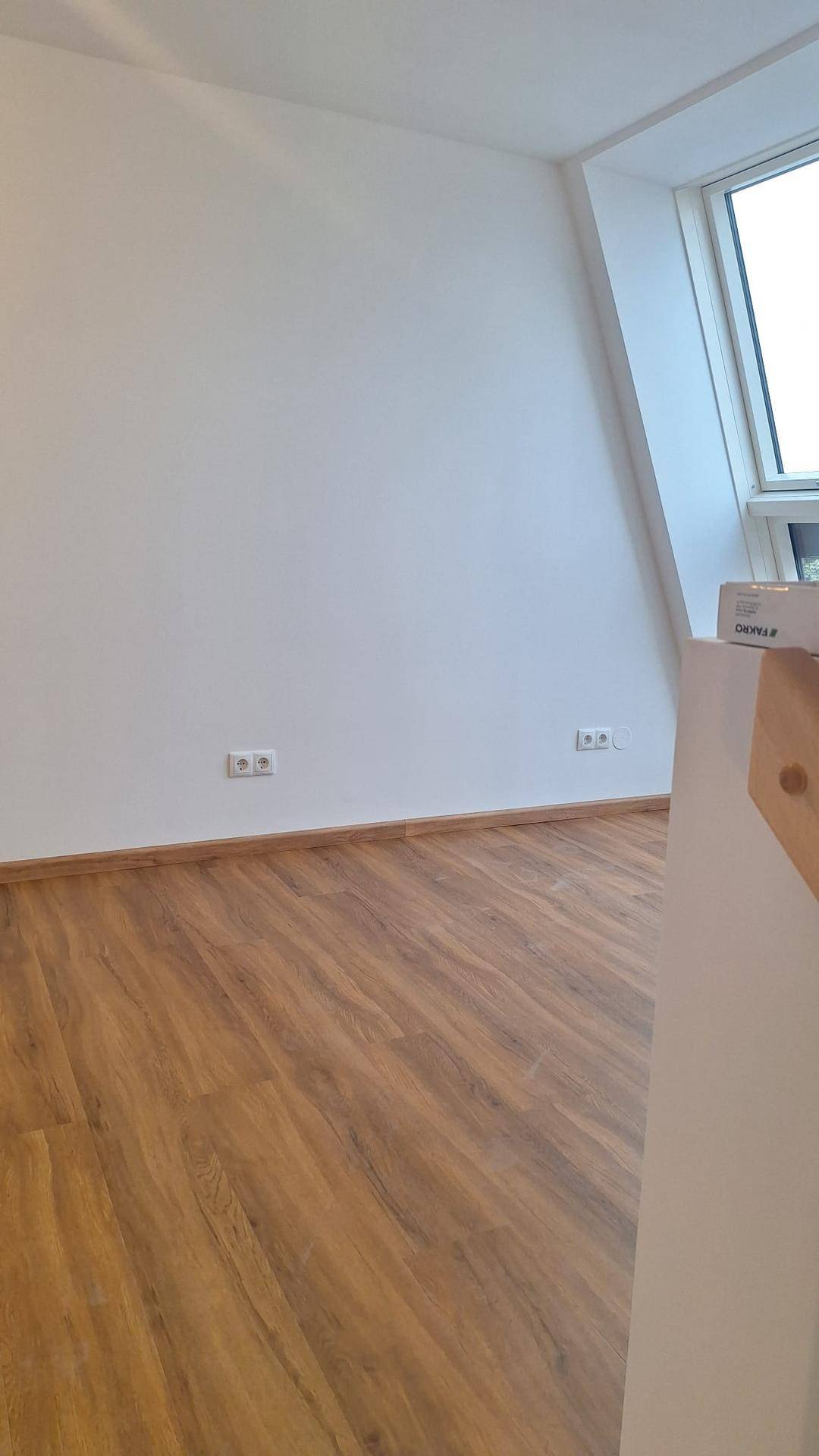 Ecke eines hellen Raumes mit Laminatboden, weißen Wänden und einem Dachfenster.