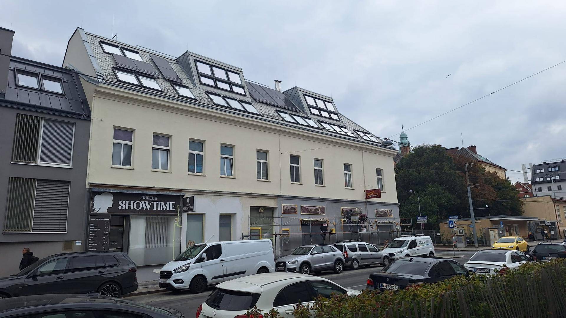 Fassade eines Gebäudes mit Mansarddach und Geschäften im Erdgeschoss, umgeben von parkenden Autos.