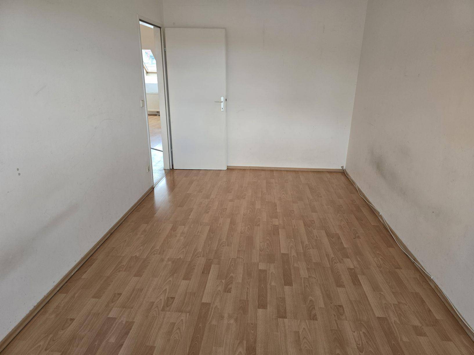 Leeres Zimmer mit Laminatboden und weiß gestrichenen Wänden, mit Zugang zu einem weiteren Raum.