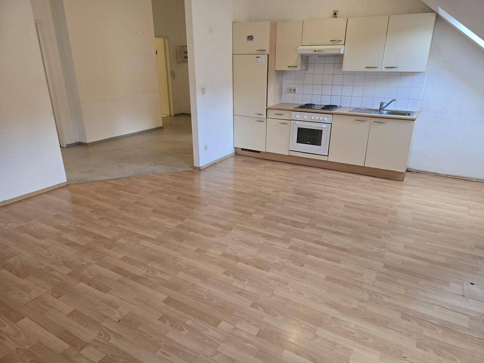 Offener Wohnbereich mit kleiner Küchenzeile und Blick in den angrenzenden Flur mit unterschiedlichem Bodenbelag.