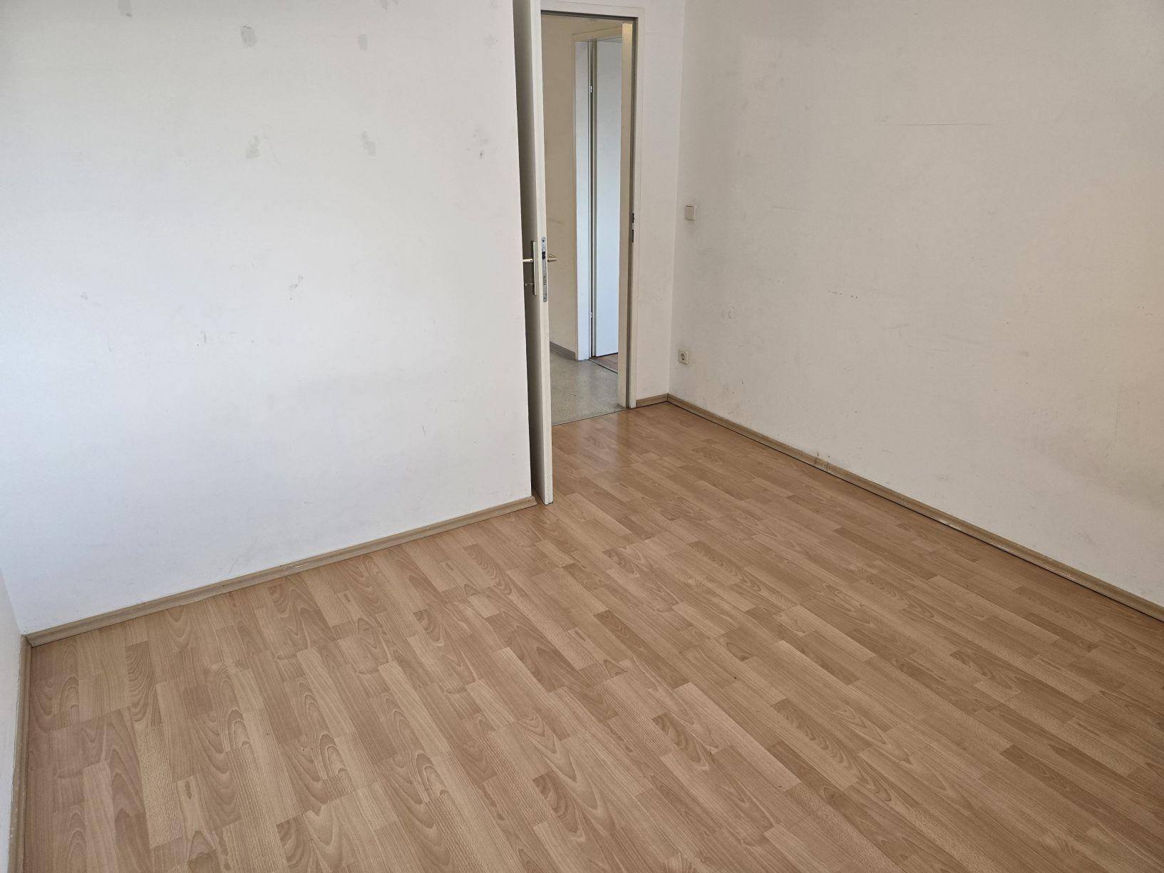 Schlichtes Zimmer mit Laminatboden und weiß gestrichenen Wänden, mit Tür zu einem angrenzenden Bereich.