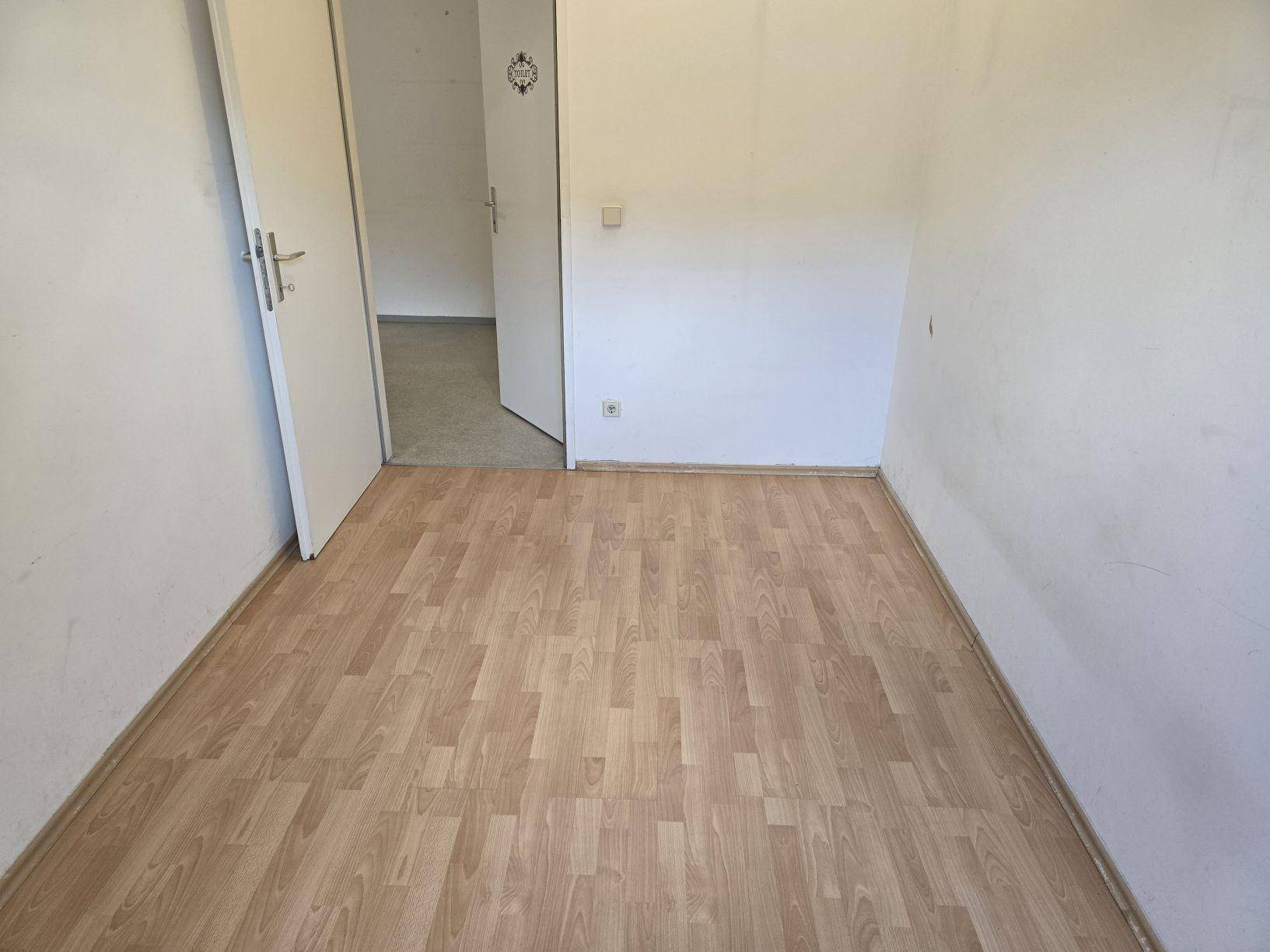 Einfaches Zimmer mit Laminatboden und weißen Wänden, mit Zugang zu einem weiteren Wohnbereich.