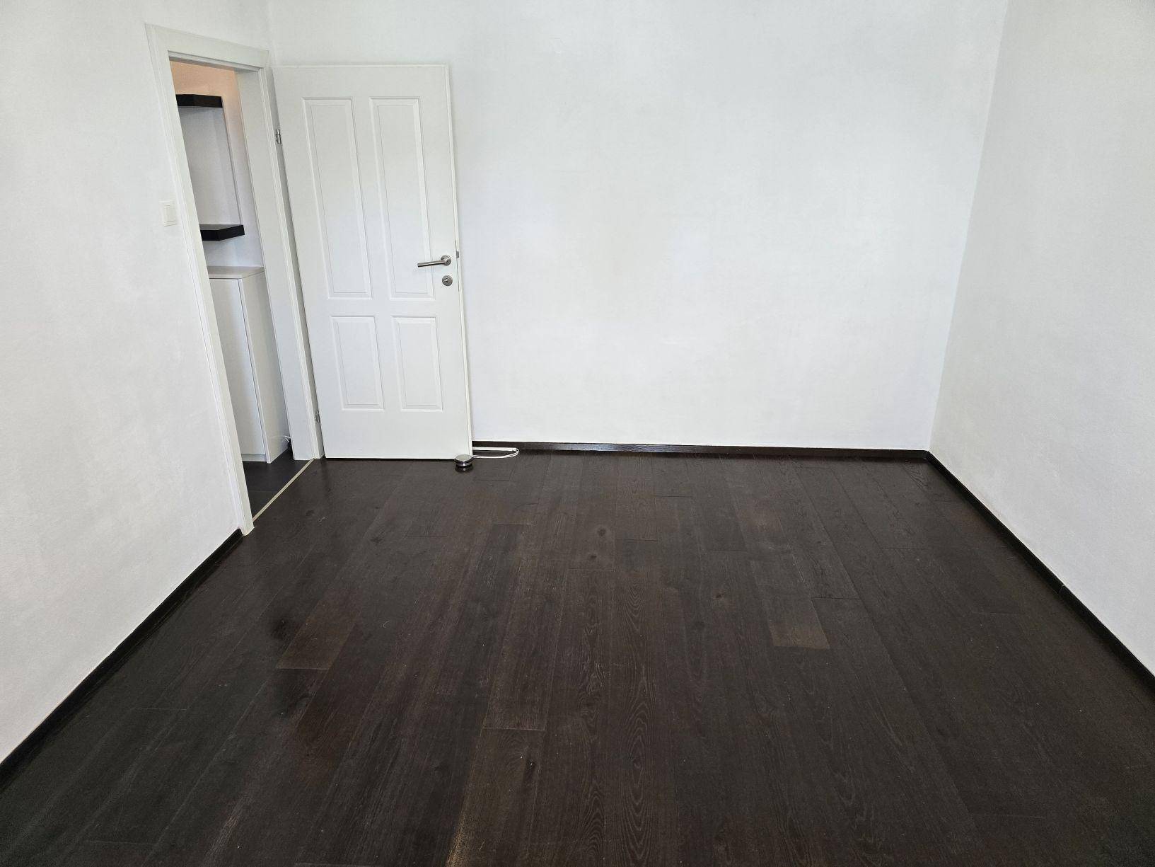 Leerer Raum mit dunklem Holzboden und einer weißen Tür, die zu einem weiteren Bereich führt.
