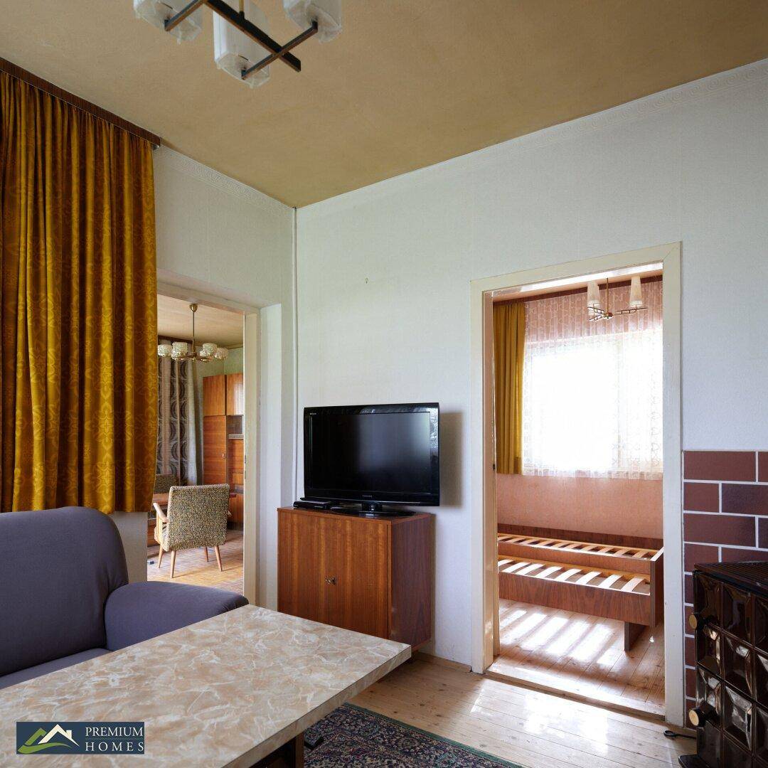 Gemütliches Wohnzimmer mit Sofa, Fernseher und traditionellem Kachelofen, durch eine Tür ist ein weiteres Zimmer sichtbar.