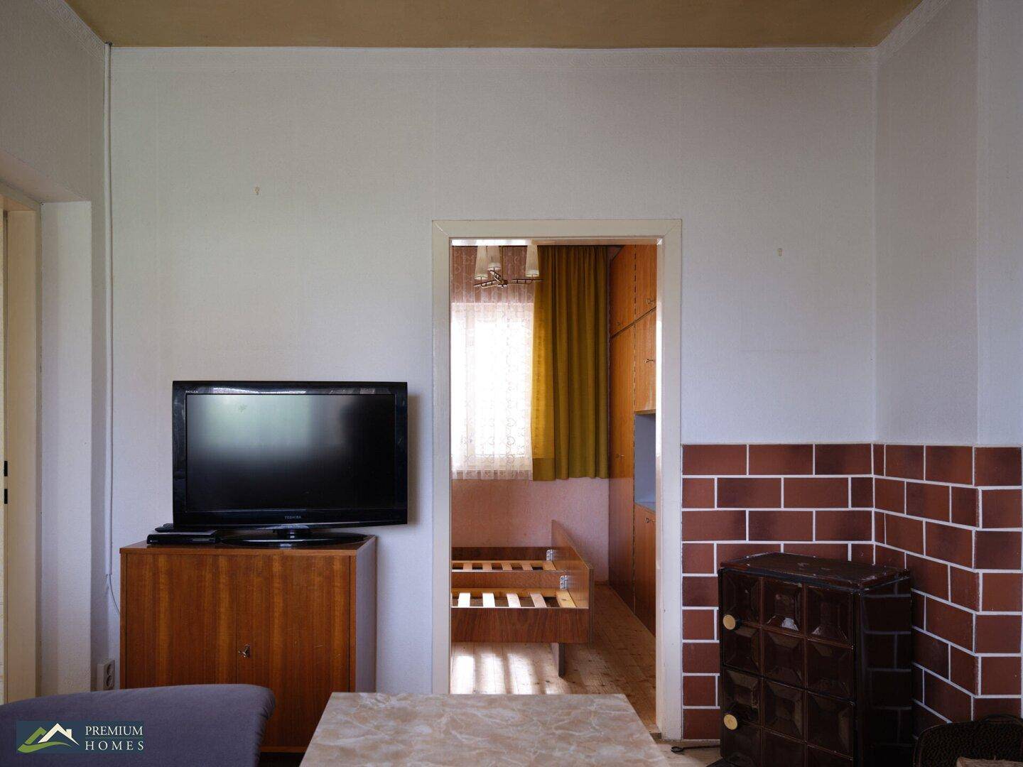 Wohnzimmer mit Fernseher, Kachelofen und Blick in ein angrenzendes Schlafzimmer mit Bettgestell.