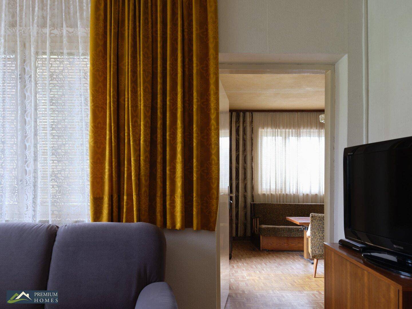 Wohnzimmer mit Sofa und Vorhängen, die einen Blick in einen weiteren Raum mit Esstisch ermöglichen.