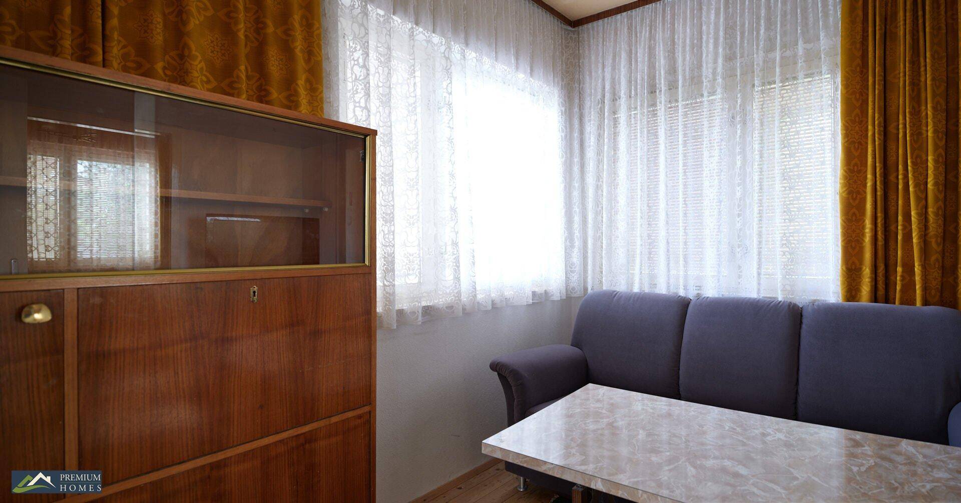 Wohnzimmer mit Sofa, Couchtisch und einem Holzschrank vor einem großen Fenster mit Vorhängen.
