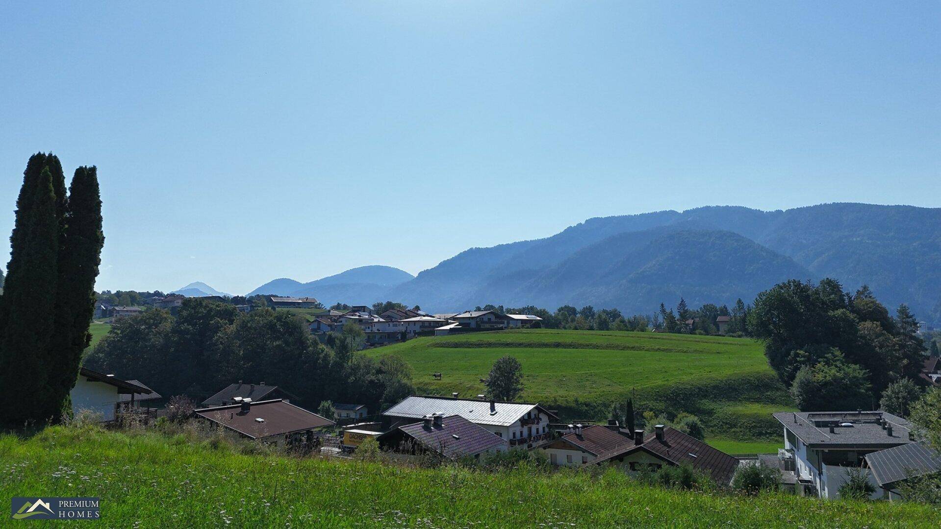 Weitläufige Landschaft mit Häusern, grünen Feldern und einer beeindruckenden Bergkulisse unter blauem Himmel.