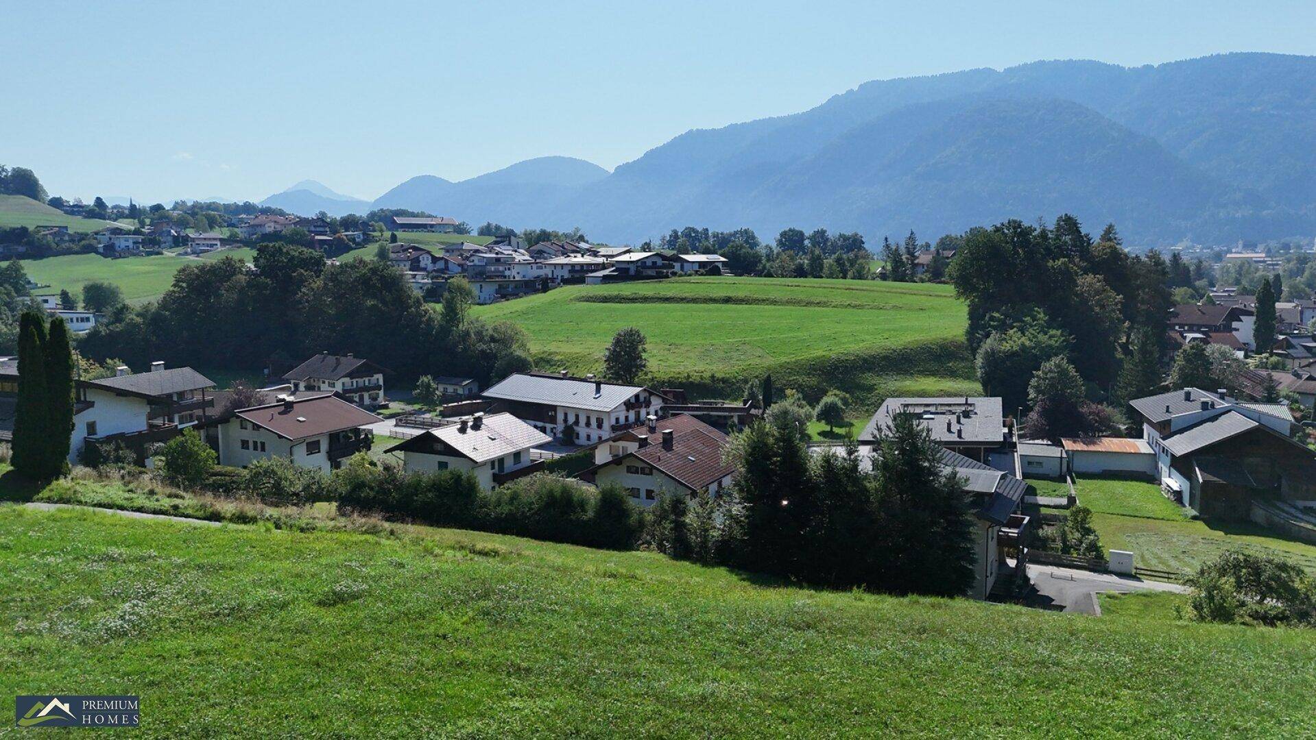 Panoramablick auf eine ländliche Siedlung mit grünen Hügeln und einer weiten Berglandschaft im Hintergrund.