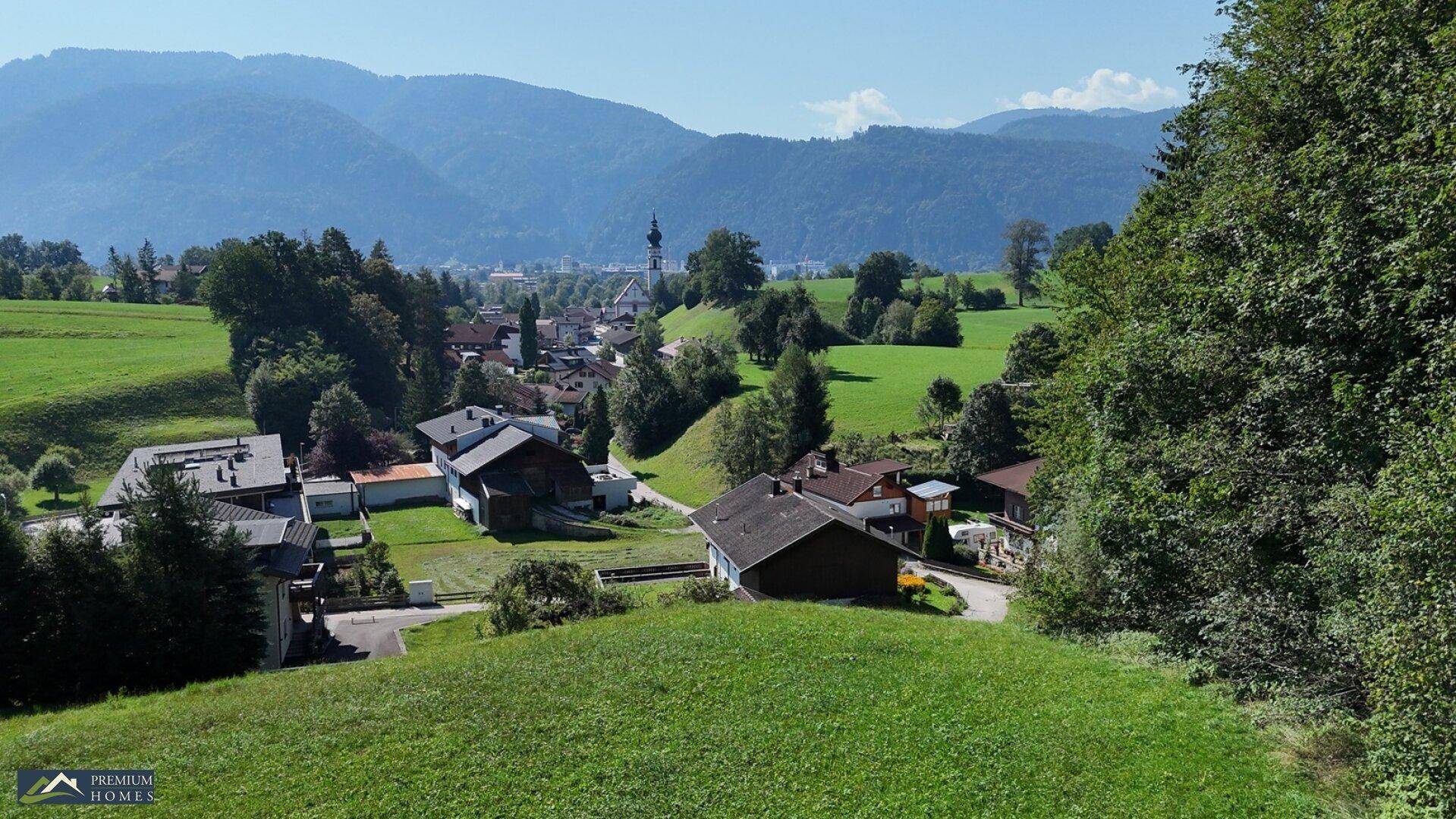 Malerischer Ausblick auf ein Dorf mit Kirchturm, eingebettet in eine grüne Hügellandschaft und Berge.