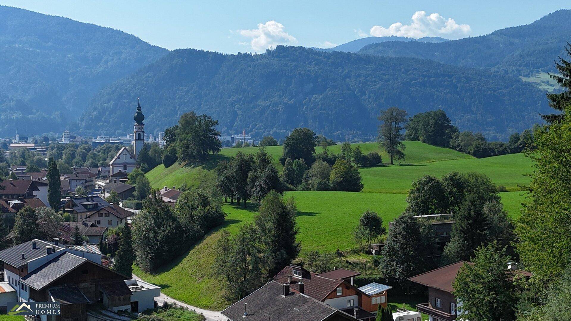 Weitwinkelaufnahme einer ländlichen Szenerie mit Häusern, Feldern und einem Kirchturm vor einer Bergkulisse.