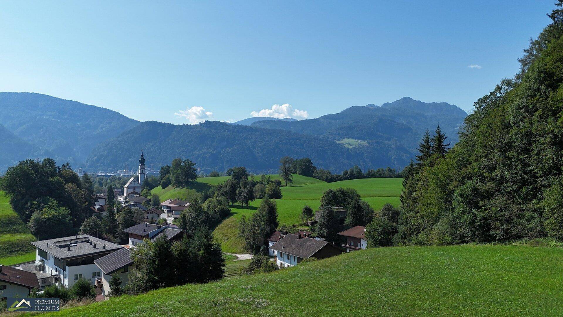 Panoramabild einer Siedlung in einer grünen Talsenke, umrahmt von bewaldeten Hügeln und hohen Bergen.