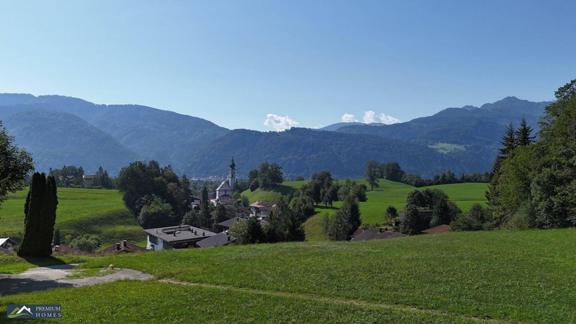 Idyllische Landschaft mit Häusern, grünen Hügeln und einem Kirchturm, umgeben von einer Bergkette.