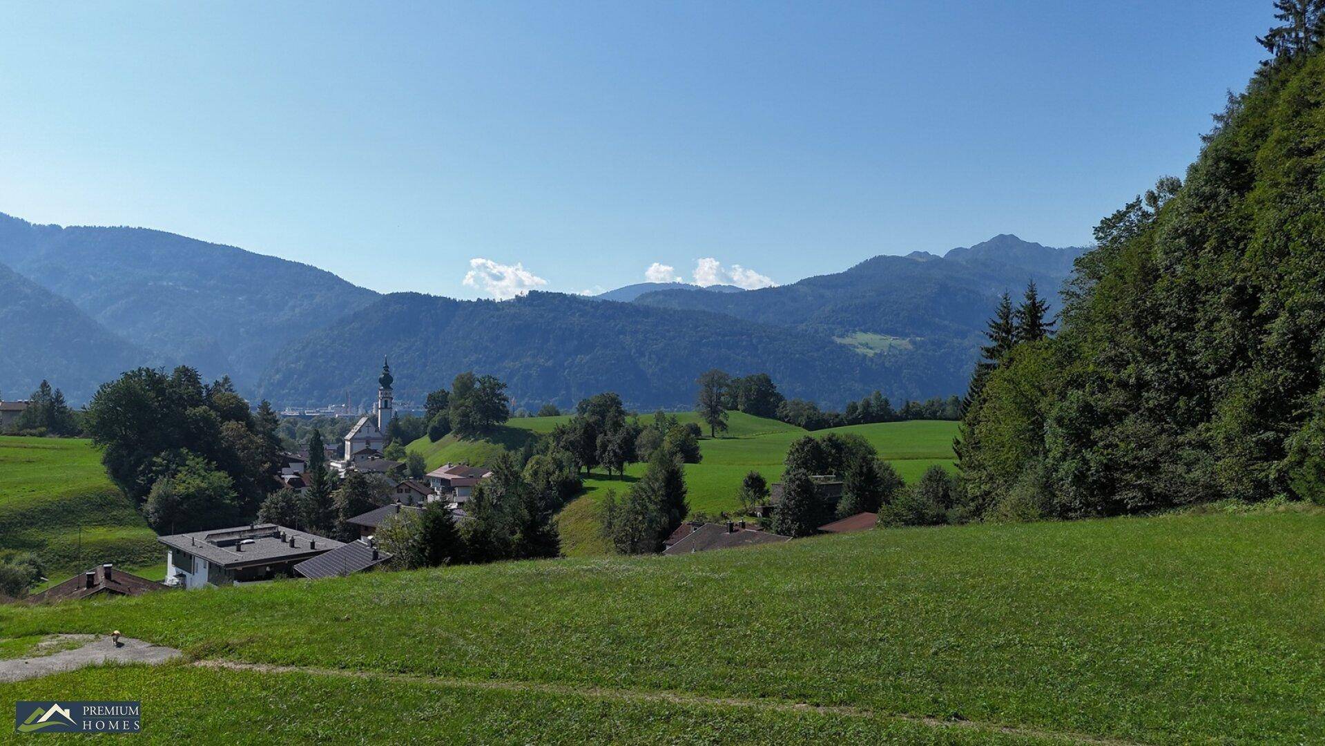 Panoramablick auf eine ländliche Gegend mit Häusern, grünen Wiesen und Bergen im Hintergrund.
