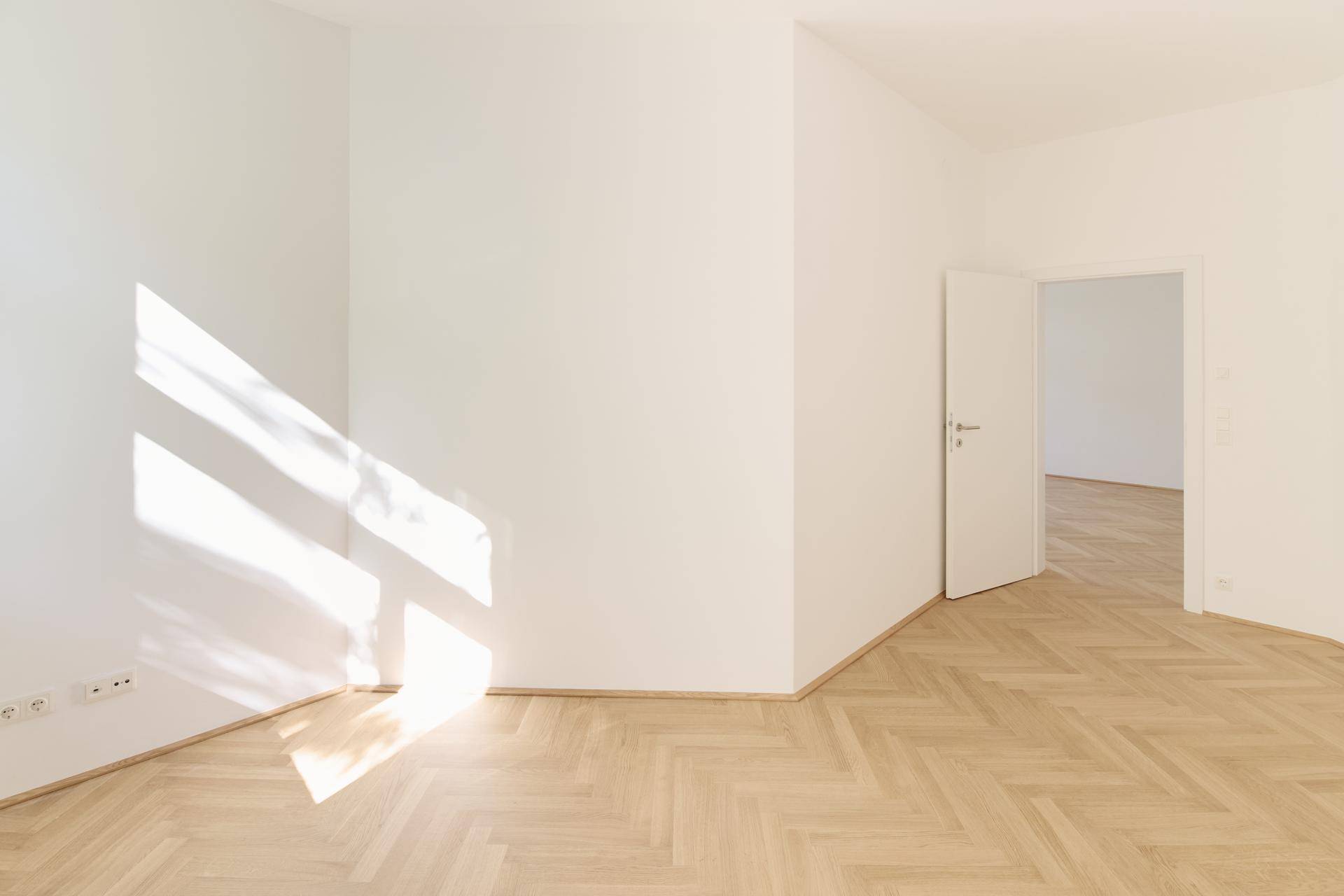 Lichtdurchfluteter Raum mit Fischgrätparkett und sonnigen Lichtstrahlen auf dem Boden.