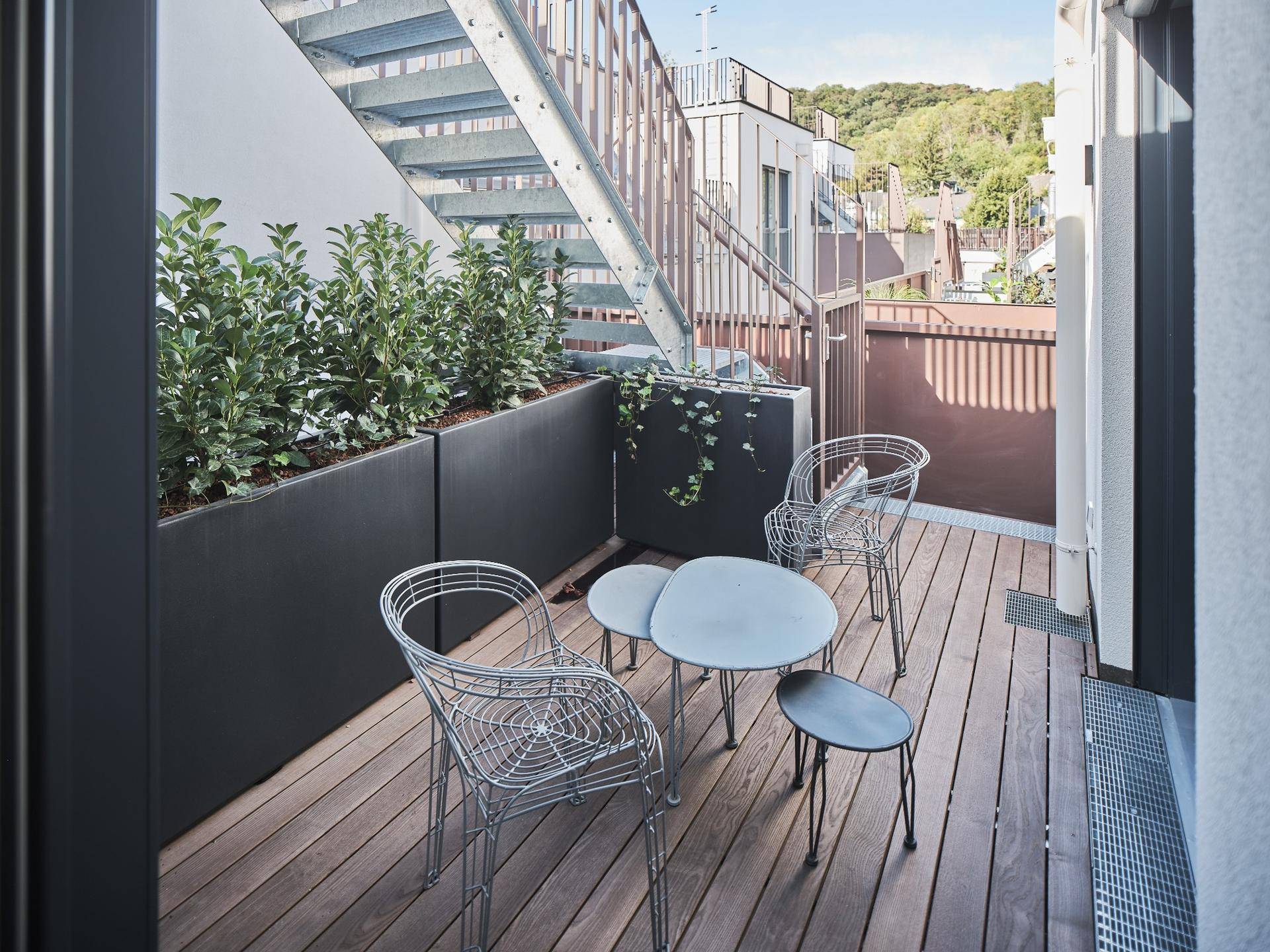 Kleiner Balkon mit Holzboden, Sitzgelegenheiten und Pflanzenkübeln, zugänglich über eine Außentreppe.
