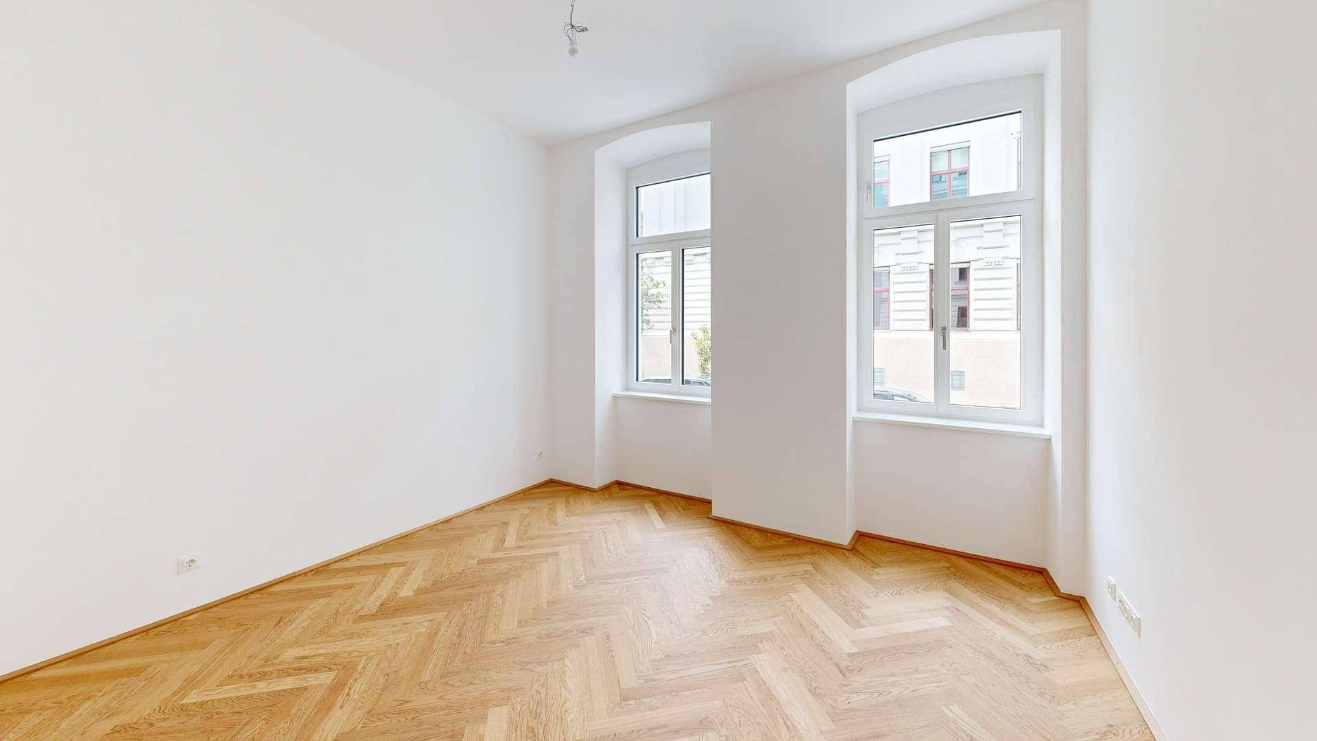 Helles Zimmer mit Fischgrätparkettboden und zwei großen Fenstern, die viel Tageslicht hereinlassen.