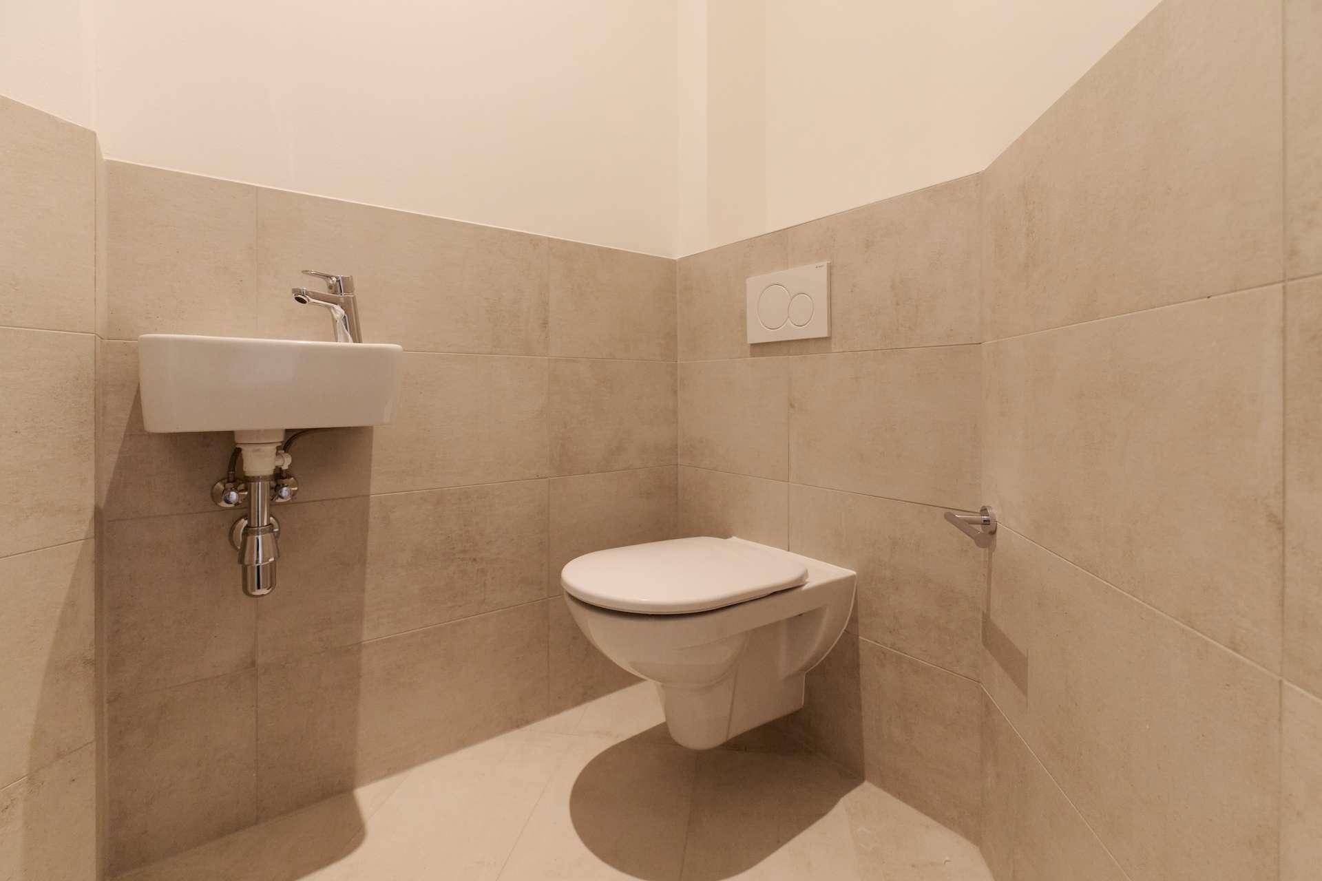 Gäste-WC mit wandhängender Toilette, kleinem Waschbecken und beigefarbenen Fliesen.