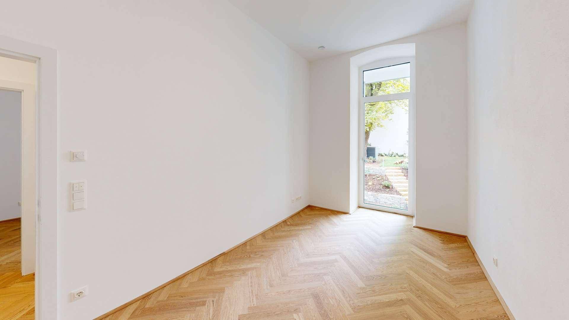 Lichtdurchflutetes Zimmer mit Fischgrätparkett und hoher Fenstertür mit Blick in den Garten.