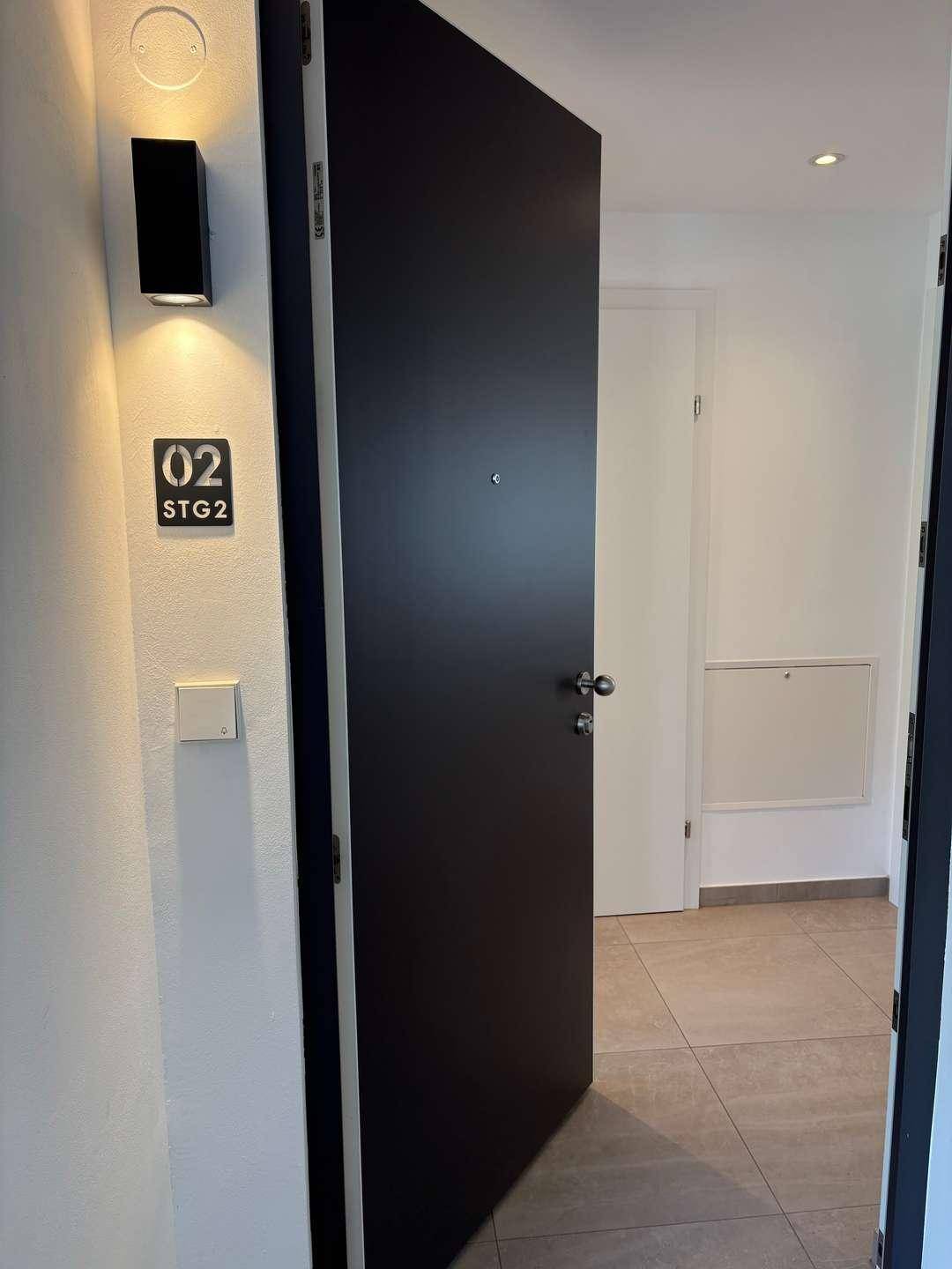 Eingangsbereich mit moderner schwarzer Tür, Wandleuchte und Fliesenboden.
