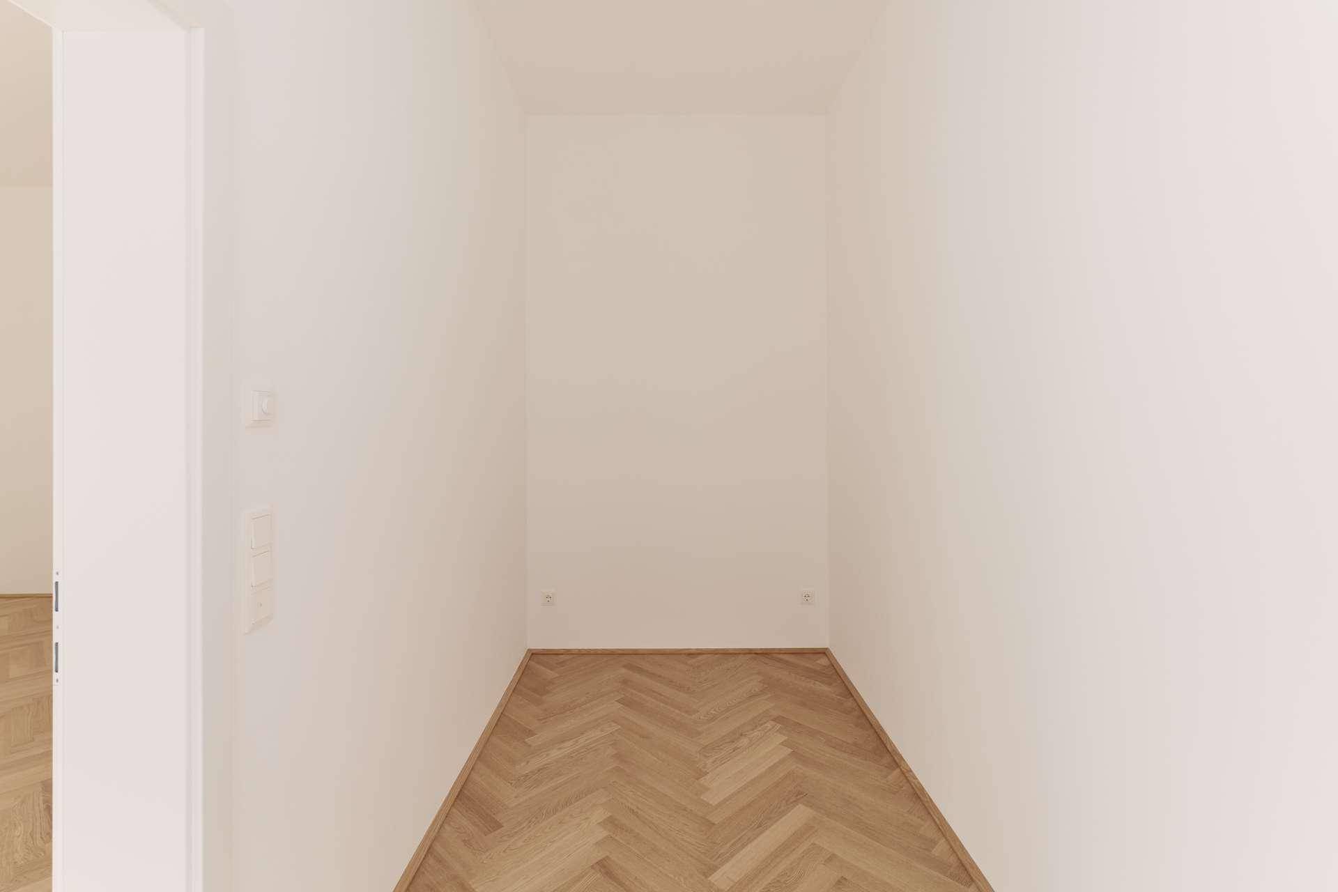 Schmaler Raum mit Fischgrätparkettboden und weißen Wänden, ideal als Abstellraum oder Ankleidezimmer.