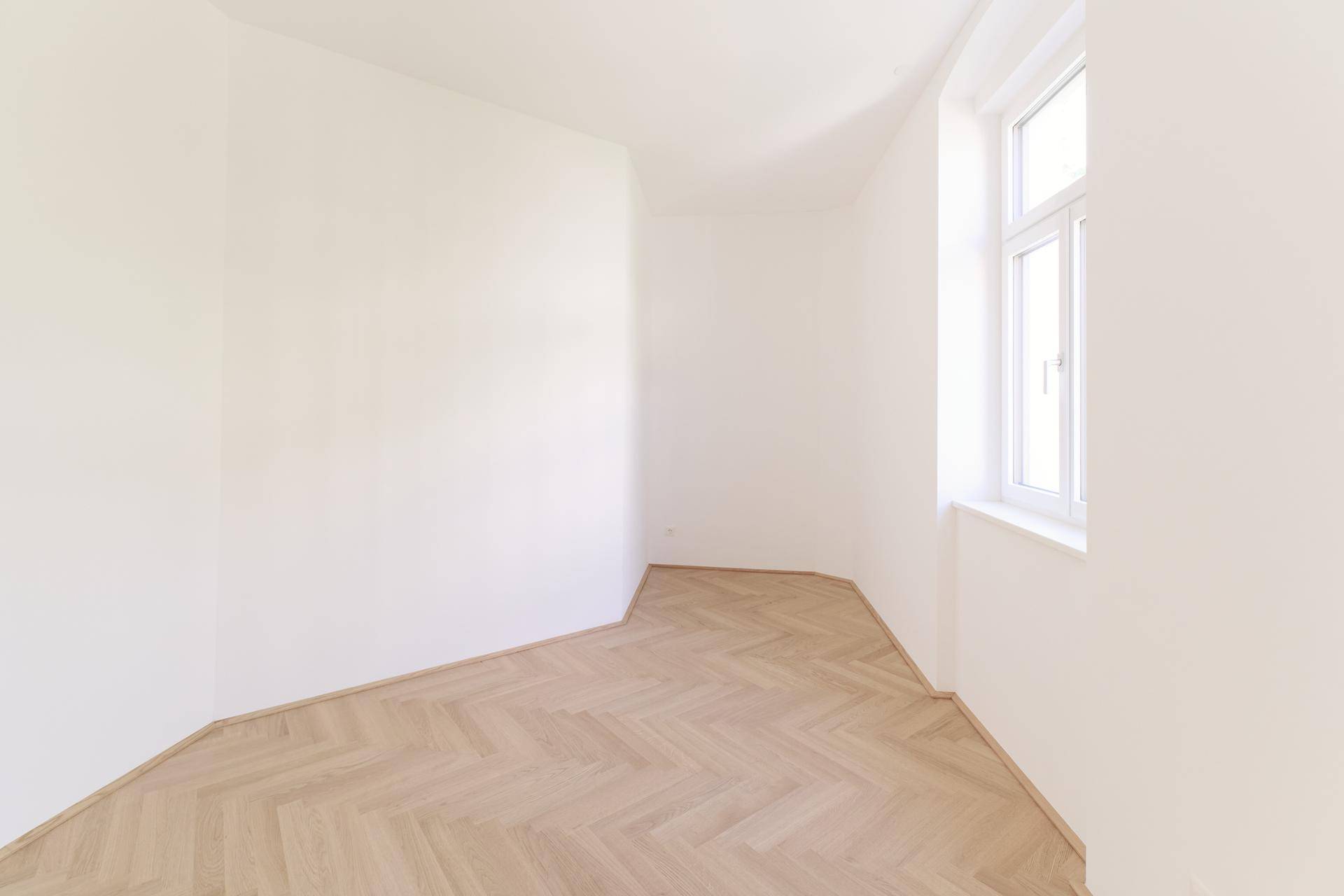Heller Raum mit Fischgrätparkett und einem Fenster, das viel Tageslicht hereinlässt.