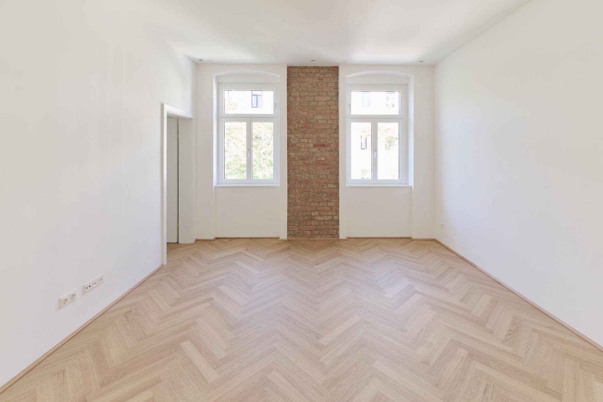 Geräumiges Zimmer mit Fischgrätparkett, zwei Fenstern und einer dekorativen Ziegelwand.