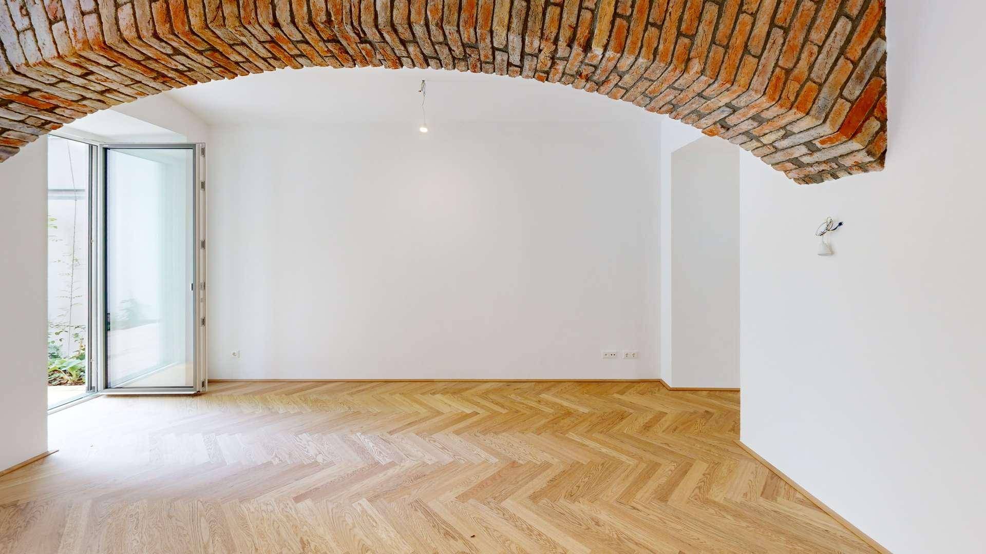 Heller Raum mit Fischgrätparkett, freigelegtem Ziegelgewölbe und Glastür zum Außenbereich.