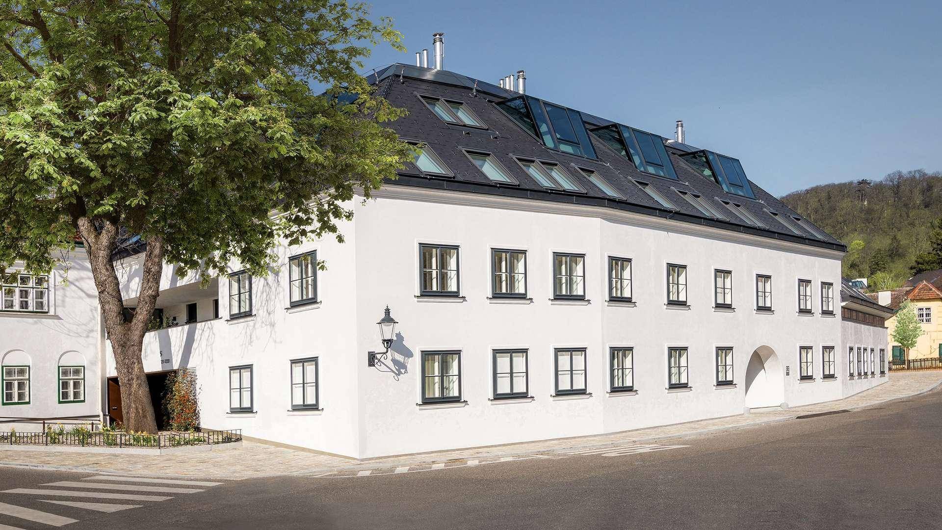 Elegantes weißes Gebäude mit dunklem Mansarddach und vielen Fenstern in grüner Umgebung.