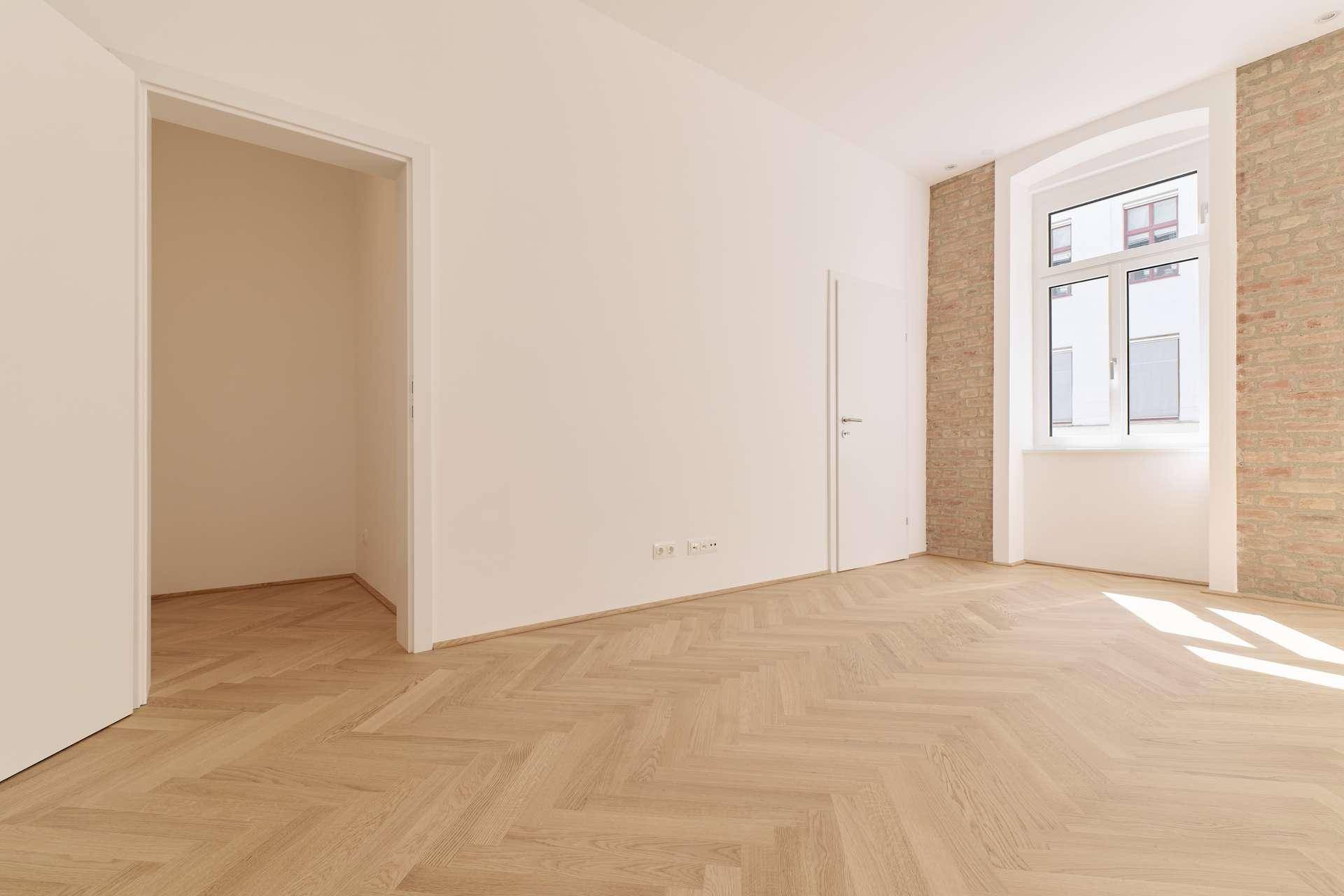 Geräumiges Zimmer mit Fischgrätparkett, weiß gestrichenen Wänden und einem Fenster mit Ziegelmauerwerk.